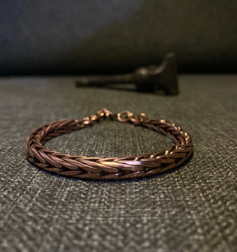 K&ouml;nnte beinhalten: Ein kupferfarbenes geflochtenes Armband mit strukturiertem Design. Das Armband besteht aus ineinandergreifenden Metallgliedern und hat einen Verschluss. Der Hintergrund ist eine dunkelgraue Oberfl&auml;che.