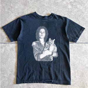 May include: Dark navy blue t-shirt featuring a black and white image of a person holding a cat. The image is a portrait of a person with curly hair and a cat. The t-shirt has a crew neck and short sleeves.