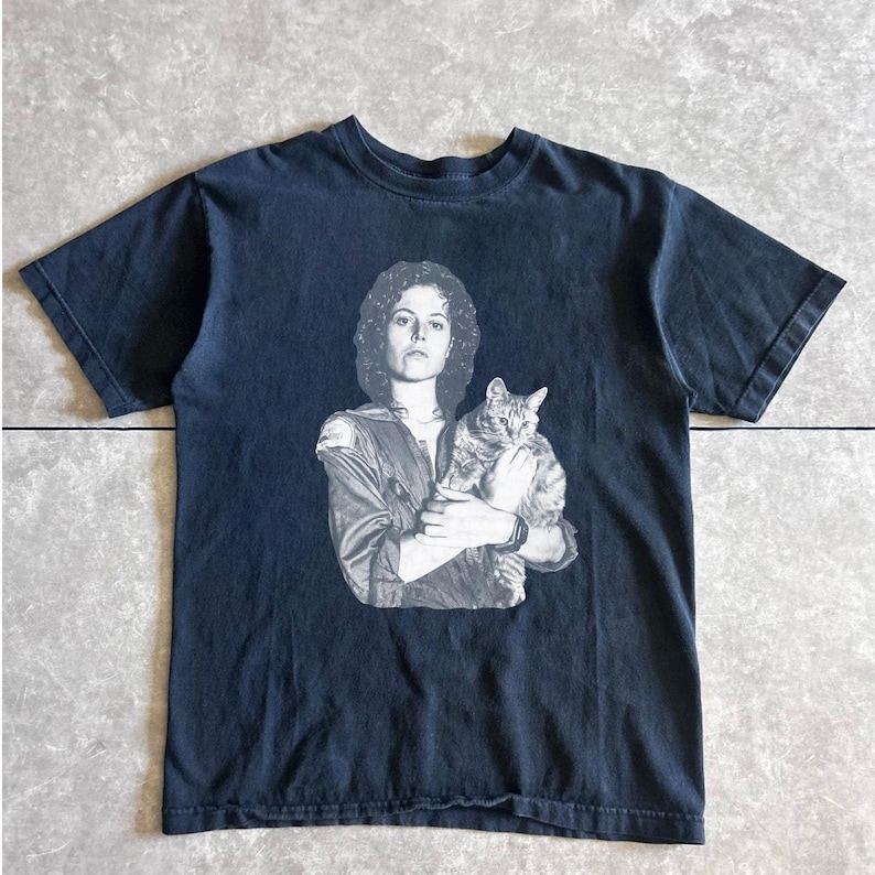 May include: Dark navy blue t-shirt featuring a black and white image of a person holding a cat. The image is a portrait of a person with curly hair and a cat. The t-shirt has a crew neck and short sleeves.