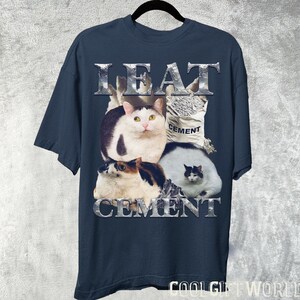 Pode incluir: Camiseta azul marinho com um gr&aacute;fico de quatro gatos e um saco de cimento. As palavras "LEAT CEMENT" s&atilde;o impressas em prata, com uma fonte texturizada acima e abaixo dos gatos. A camiseta est&aacute; pendurada em um cabide preto.