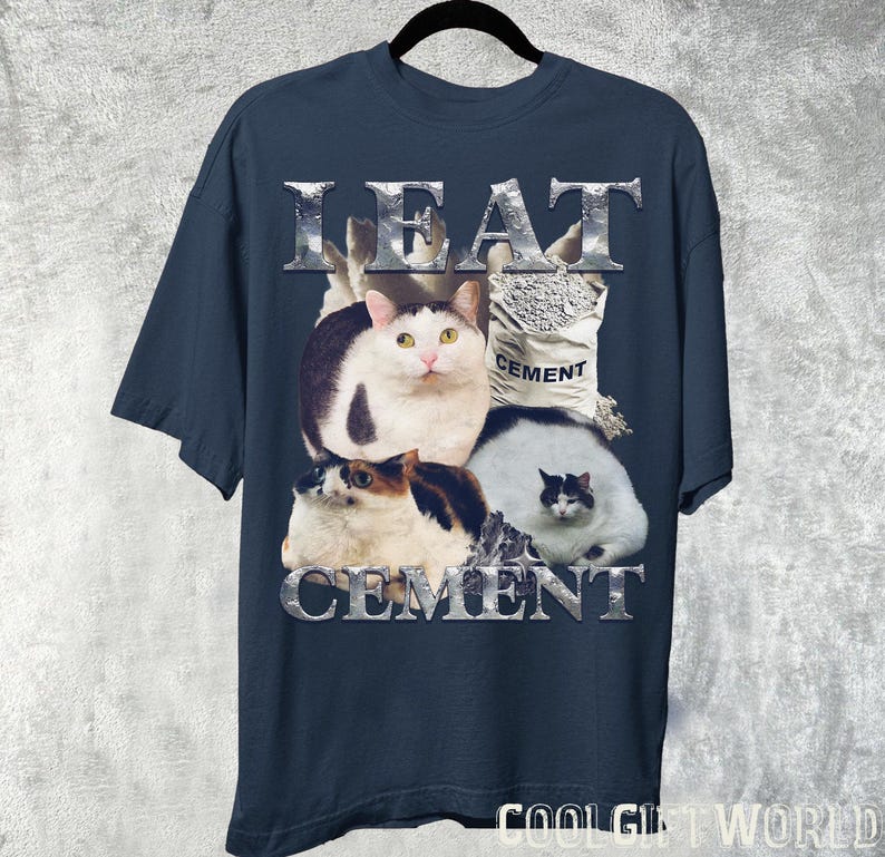 Pode incluir: Camiseta azul marinho com um gr&aacute;fico de quatro gatos e um saco de cimento. As palavras "LEAT CEMENT" s&atilde;o impressas em prata, com uma fonte texturizada acima e abaixo dos gatos. A camiseta est&aacute; pendurada em um cabide preto.