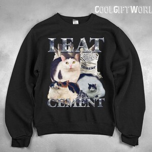 Pode incluir: Camisola preta com um gr&aacute;fico de quatro gatos e um saco de cimento. O texto "EAT CEMENT" &eacute; exibido em letras met&aacute;licas acima e abaixo dos gatos. A camisola tem gola redonda e mangas compridas.