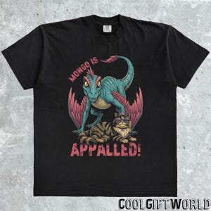 May include: Black t-shirt featuring a cartoon illustration of a blue and red dinosaur with a cat wearing sunglasses. The text "MONGO IS APPALLED!" is printed in pink.
