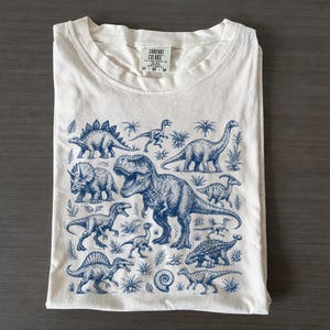 May include: A cream-colored short-sleeve t-shirt featuring a detailed blue ink-style graphic print of various dinosaurs and prehistoric plants. The inner neck label displays the text Comfort Colors and M-M, indicating the size of the garment.