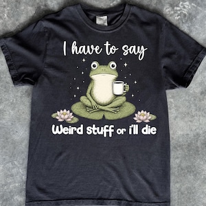 May include: A black short-sleeve t-shirt featuring a graphic of a green frog sitting on a lily pad holding a white mug. The text reads I have to say Weird stuff or i'll die. Two pink lotus flowers are positioned on either side of the lily pad.