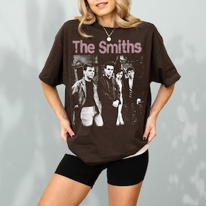 May include: A brown t-shirt featuring the band "The Smiths" with a black and white image of the band members. The band's name is printed in a light purple color across the top of the image.