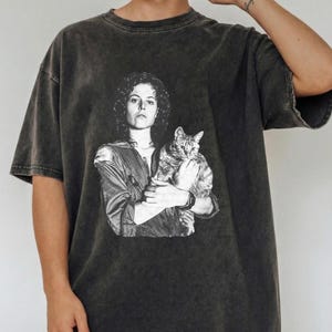 May include: Dark gray t-shirt featuring a black and white graphic of a woman holding a cat. The image is a vintage-style print on a short-sleeved, oversized tee. The shirt has a faded, worn look.