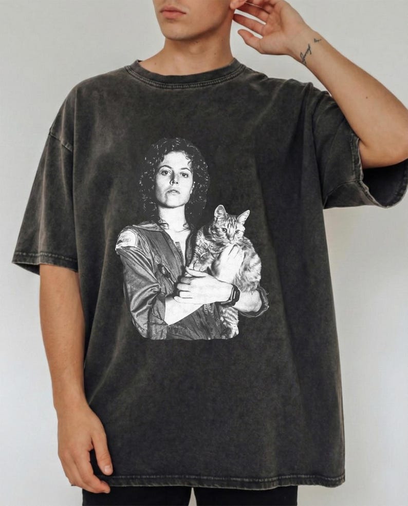 May include: Dark gray t-shirt featuring a black and white graphic of a woman holding a cat. The image is a vintage-style print on a short-sleeved, oversized tee. The shirt has a faded, worn look.