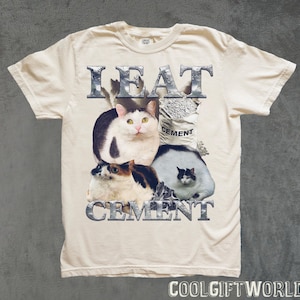 Pode incluir: Camiseta creme com um gr&aacute;fico de tr&ecirc;s gatos e um saco de cimento. O texto "I EAT CEMENT" &eacute; impresso acima e abaixo dos gatos em uma fonte met&aacute;lica. A camiseta tem mangas curtas.