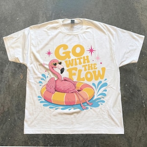 May include: A white short-sleeve t-shirt featuring a graphic of a pink flamingo wearing heart-shaped sunglasses, floating on a striped orange and yellow inner tube. The text GO WITH THE FLOW is printed in yellow above the flamingo with pink stars.