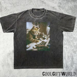 May include: Dark gray t-shirt featuring a graphic of two cats, one grooming the other. The image is in a rectangular frame. The text "COOLGIFTWORLD" is printed in white at the bottom of the shirt.