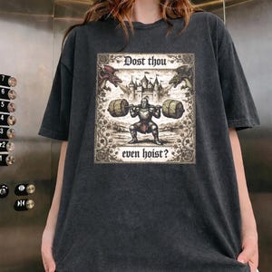 May include: Dark grey t-shirt with a medieval-themed graphic. The graphic shows a knight lifting weights, dragons, a castle, and the text "Dost thou even hoist?" The shirt has a vintage, worn appearance.
