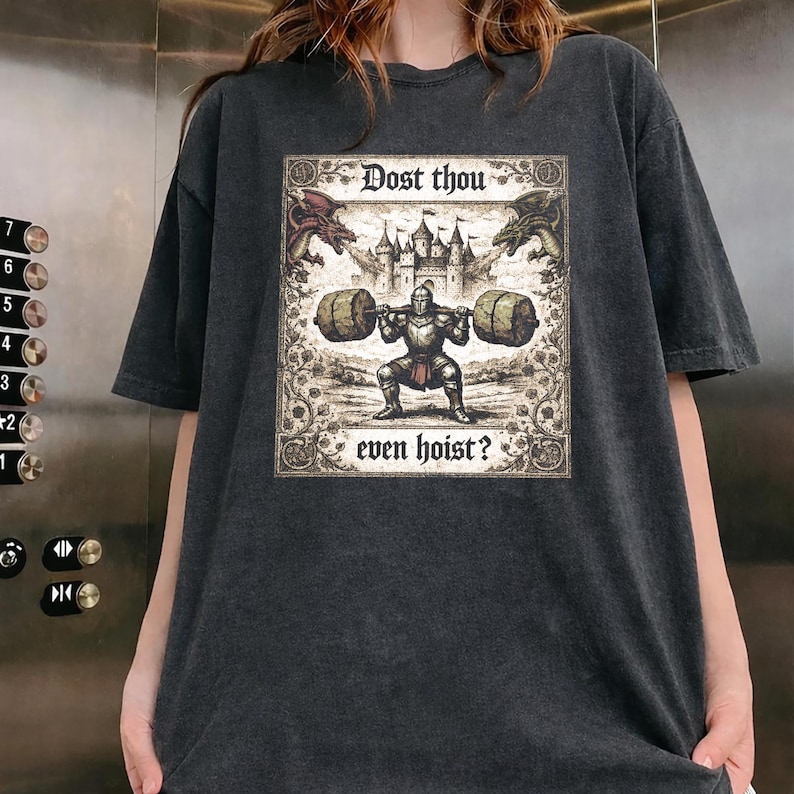 May include: Dark grey t-shirt with a medieval-themed graphic. The graphic shows a knight lifting weights, dragons, a castle, and the text "Dost thou even hoist?" The shirt has a vintage, worn appearance.