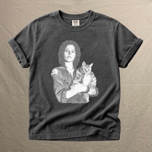 May include: Dark gray t-shirt featuring a black and white image of a person holding a cat. The image is a vintage style print. The shirt has rolled-up sleeves and a crew neck. The tag reads "COMFORT COLORS".