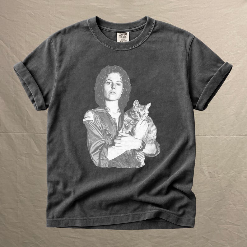 May include: Dark gray t-shirt featuring a black and white image of a person holding a cat. The image is a vintage style print. The shirt has rolled-up sleeves and a crew neck. The tag reads "COMFORT COLORS".