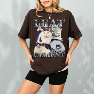 Pode incluir: Uma camiseta marrom com um gr&aacute;fico de tr&ecirc;s gatos e um saco de cimento. O texto "I EAT CEMENT" &eacute; impresso acima e abaixo do gr&aacute;fico do gato em uma fonte met&aacute;lica. A camiseta est&aacute; sendo usada.