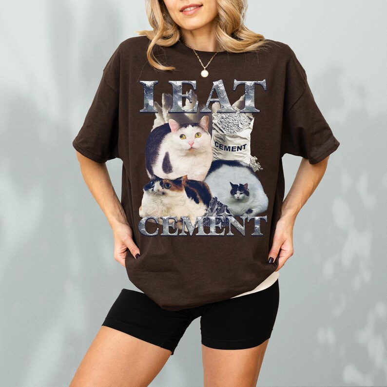 Pode incluir: Uma camiseta marrom com um gr&aacute;fico de tr&ecirc;s gatos e um saco de cimento. O texto "I EAT CEMENT" &eacute; impresso acima e abaixo do gr&aacute;fico do gato em uma fonte met&aacute;lica. A camiseta est&aacute; sendo usada.