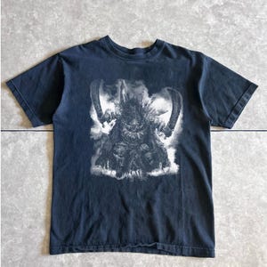 May include: Dark blue t-shirt featuring a detailed black and white graphic of a creature with large curved blades. The design is centered on the shirt, with the creature appearing to be in a battle stance. The shirt has short sleeves and a crew neck.