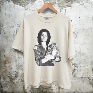 May include: Cream-colored t-shirt featuring a black and white graphic of a person holding a cat. The person is wearing a jacket. The t-shirt is hanging on a wooden hanger.