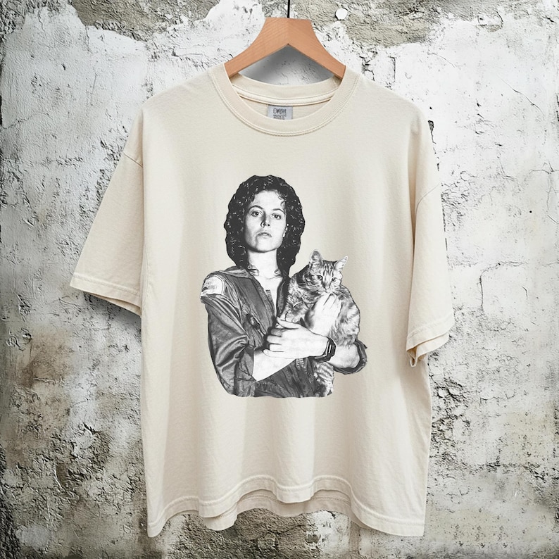May include: Cream-colored t-shirt featuring a black and white graphic of a person holding a cat. The person is wearing a jacket. The t-shirt is hanging on a wooden hanger.
