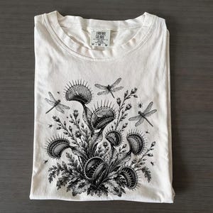 Może przedstawiać: Kremowy t-shirt z krótkim rękawem z czarną ilustracją w stylu tuszu przedstawiającą kilka muchołówek i ważek. Wewnętrzna metka na karku zawiera napis Comfort Colors oraz rozmiar M. Wzór zawiera szczegółowe elementy botaniczne.