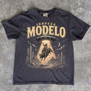 May include: A charcoal gray t-shirt featuring a graphic print of a cowboy riding a bull being lifted by a UFO. The text CERVEZA MODELO is printed in large beige letters above the saucer. The scene includes desert cacti and a crescent moon.