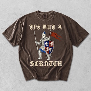 May include: A brown short-sleeve t-shirt featuring a graphic of a medieval knight in chainmail armor holding a spear and a shield with fleur-de-lis and lion emblems. A red flag waves behind the knight. The text TIS BUT A SCRATCH is printed.