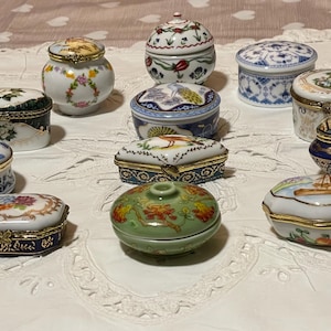 Pode incluir: Uma coleção de caixas de joias decorativas de porcelana em várias formas e tamanhos. As caixas apresentam desenhos florais, paisagísticos e geométricos com acabamentos dourados. Algumas têm tampas com pináculos ornamentados, e a paleta de cores inclui branco, azul, verde e dourado.
