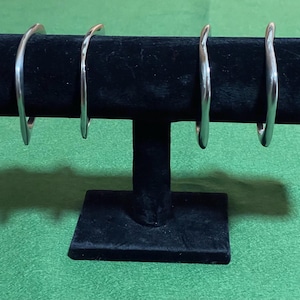 Può includere: Quattro braccialetti argentati e curvi esposti su un espositore di gioielli in velluto nero. I braccialetti hanno una finitura liscia e lucida e un design semplice ed elegante. L'espositore è su una superficie verde, creando un contrasto.
