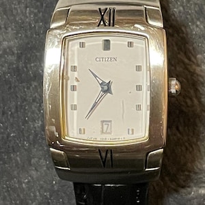 Könnte beinhalten: Eine silberne Citizen-Armbanduhr mit rechteckigem Zifferblatt und schwarzem Lederarmband. Das Zifferblatt ist weiß mit schwarzen römischen Ziffern und Zeigern. Das Datum wird bei 3 Uhr angezeigt. Klassisches Uhrendesign.