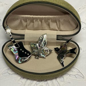 May include: An open olive green jewellery case displaying three decorative brooches. The brooches include a guitar, a leaf, and a bird, all with iridescent abalone shell inlays. The case has a beige interior and a zip closure.