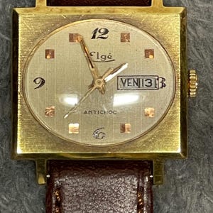May include: A vintage gold-toned Elgé wristwatch with a square face and a brown leather strap. The watch face is light gold with gold hands and markers. The date reads "VEN 13".