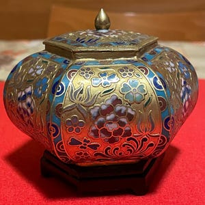 Pode incluir: Um recipiente ornamentado com tampa, com base e tampa em metal dourado. Apresenta intrincados desenhos florais em tons de azul, branco e vermelho sobre um fundo dourado. Assenta num pequeno suporte de madeira preta.