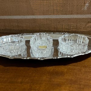 Può includere: Un vassoio ovale argentato con tre ciotole di vetro tagliato. Le ciotole sono quadrate con bordi arrotondati e un motivo decorativo. Il vassoio ha un bordo smerlato e poggia su una superficie di legno.