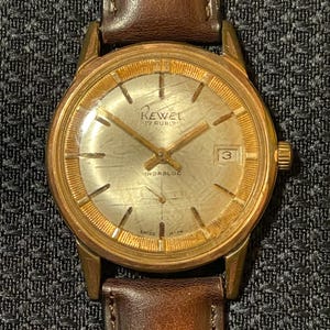 Può includere: Un orologio Rewel vintage color oro con cinturino in pelle marrone. Il quadrante dell'orologio ha una superficie strutturata di colore chiaro con lancette e indici dorati. L'orologio mostra la data e il testo "17 Rubis" e "Swiss Made".