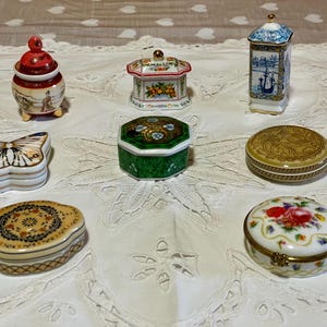 Pode incluir: Uma coleção de caixas de bugigangas decorativas de porcelana em várias formas e designs. As caixas apresentam obras de arte detalhadas, incluindo padrões florais, borboletas e ilustrações cênicas. As cores incluem branco, dourado, vermelho, verde e azul. As caixas estão dispostas sobre uma toalha de mesa de renda branca.