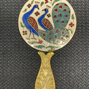 May include: A decorative hand mirror with a round, cream-coloured face featuring two blue peacocks with red and brown accents. The handle is gold-coloured with an ornate peacock design. The mirror's face is approximately 7.6 cm in diameter.