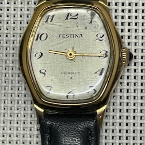 May include: A vintage Festina wristwatch with a gold-toned case and a black leather strap. The watch face is off-white with black numerals and gold hands. The watch has the text "Festina" and "Incabloc" on the face.