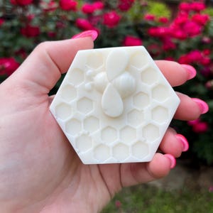May include: A white, hexagonal soap bar with a bee and honeycomb design. The bee is in the center, with a detailed honeycomb pattern covering the rest of the bar. The soap is held in a hand, with a blurred background of pink flowers.