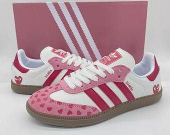 Könnte beinhalten: Weiße und rosa Adidas Samba Sneaker mit einem Herzmuster auf Zehen und Ferse. Die Sneaker haben rote Streifen und Akzente, eine braune Sohle und das Wort "Samba" auf der Seite. Im Hintergrund befindet sich eine rosa Box mit weißen Streifen.