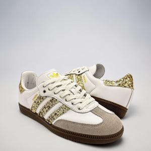 Peut inclure: Baskets en cuir blanc avec des bandes et des empiècements au talon à paillettes dorées. Les chaussures présentent un renfort en daim gris clair sur le bout, des lacets blancs et une semelle en gomme marron foncé. SAMBA est inscrit sur le côté.