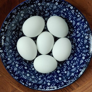 May include: Six white eggs arranged on a blue and white floral patterned plate. The plate is set inside a wooden bowl. The text "Mary's Eggs" is in the bottom right corner.