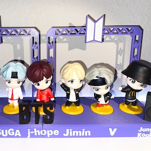 May include: A collection of seven figurines of the K-pop group BTS, each with unique hairstyles and outfits, standing on a purple base. The base has the band's name and member names. Two speakers flank the figurines.