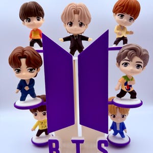 May include: A collection of seven miniature figurines of cartoon characters, each with unique hairstyles and outfits, arranged around a purple and white BTS logo. The base of the logo spells out "BTS" in white letters. The figurines are on small, white platforms.