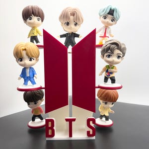 May include: A display featuring miniature figurines of seven characters, each with unique hairstyles and outfits, arranged around a red and white BTS logo. The figurines stand on small platforms, creating a layered effect.