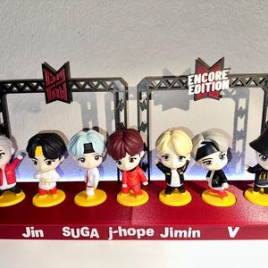 May include: A collection of seven Tiny Tan figurines, each with unique hairstyles and outfits, standing on a red base. The figurines are arranged in a row, with the names RM, Jin, SUGA, j-hope, Jimin, V, and Jung Kook printed on the base.