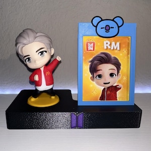 May include: A collectible figurine of a cartoon character wearing a red jacket and black pants, standing on a yellow base. Beside it is a blue frame with a cartoon image and the letters "RM". The base is black with a purple logo.