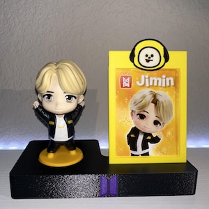 May include: A figurine of a person with blonde hair, wearing a black jacket and black pants, stands on a yellow base. A yellow card with the name "Jimin" and a cartoon character is next to the figurine. The base is black.