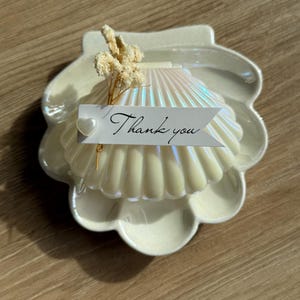 Op de afbeelding: Een parelmoerwitte schelpvormige kaars met een bijpassend schaaltje. Een klein takje gedroogde bloemen en een label met de tekst "Thank you" zijn aan de kaars bevestigd. De kaars en het schaaltje staan op een houten oppervlak.