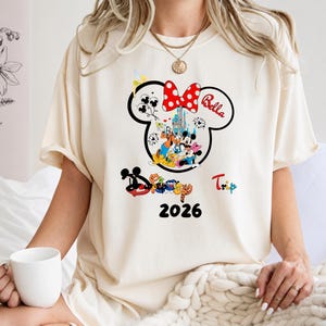 K&ouml;nnte beinhalten: Cremefarbenes T-Shirt mit einem Minnie-Mouse-Kopf-Design mit roter Schleife mit wei&szlig;en Punkten. Das Design zeigt Disney-Figuren, den Namen "Bella", das Wort "Trip" und das Jahr "2026".